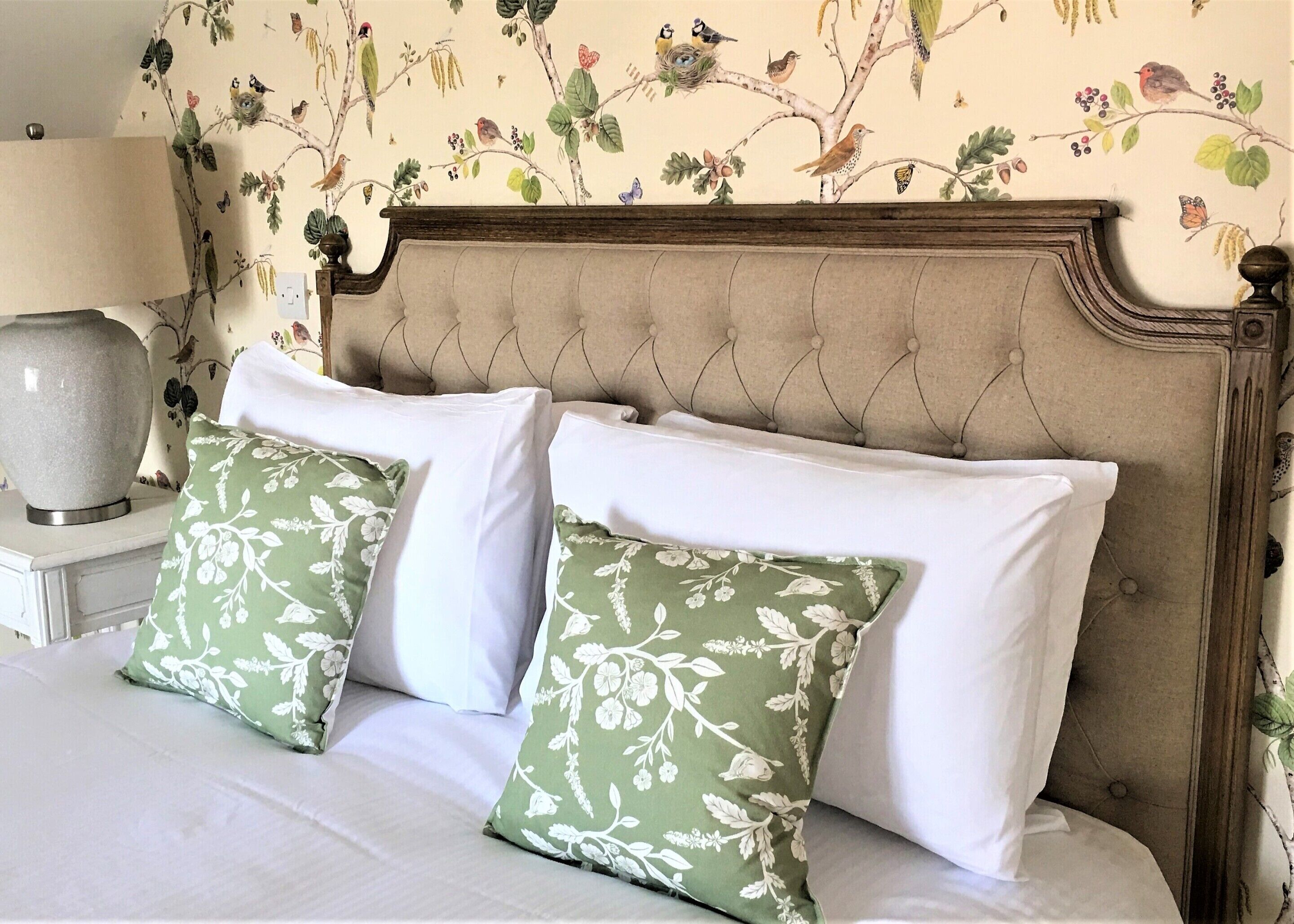 A bed with two green floral pillows and white sheets, a wooden headboard, and a nightstand with a lamp and a vase.