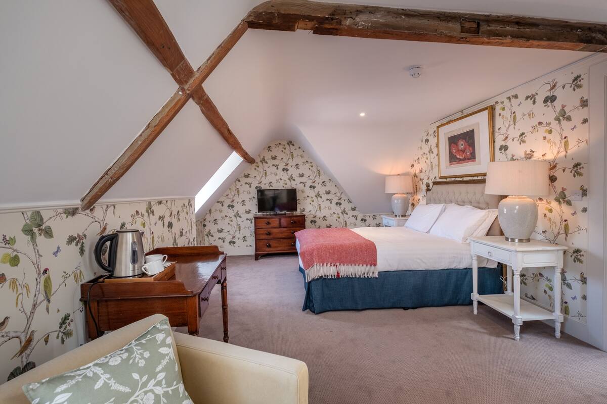 A cozy bedroom with a sloped ceiling, floral wallpaper, a bed with a blue skirt, a bedside table with a lamp, and a wooden dresser.