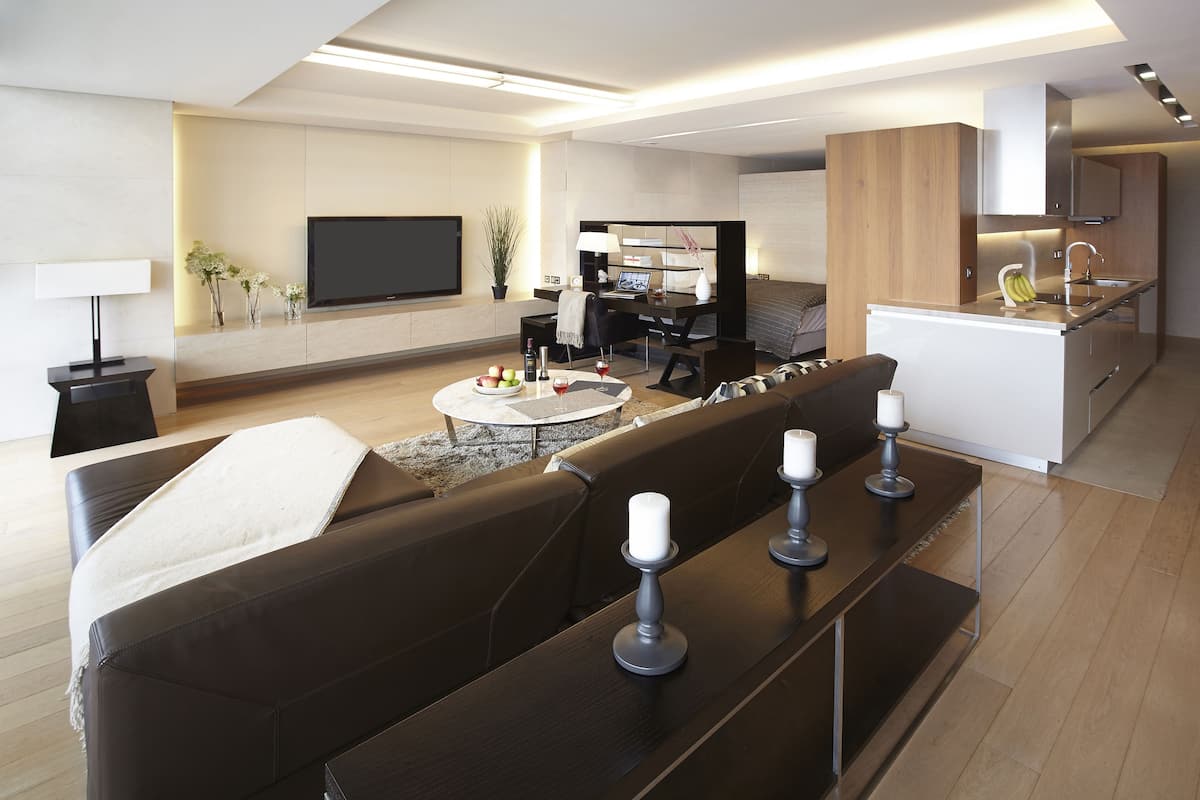 A modern living room with a sofa, a coffee table, and a TV. There is a kitchen area with a sink and cabinets.