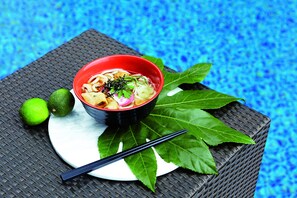 A bowl of noodle soup with lime and green leaves on a plate, placed on a woven mat.
