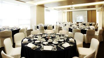 Un salón de conferencias con varias mesas redondas dispuestas para comer, cada una rodeada de sillas blancas.