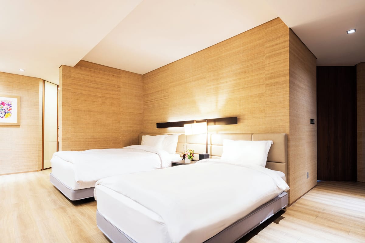 A hotel room with two beds, wooden walls, a nightstand with a lamp, and a small table with a vase of flowers.