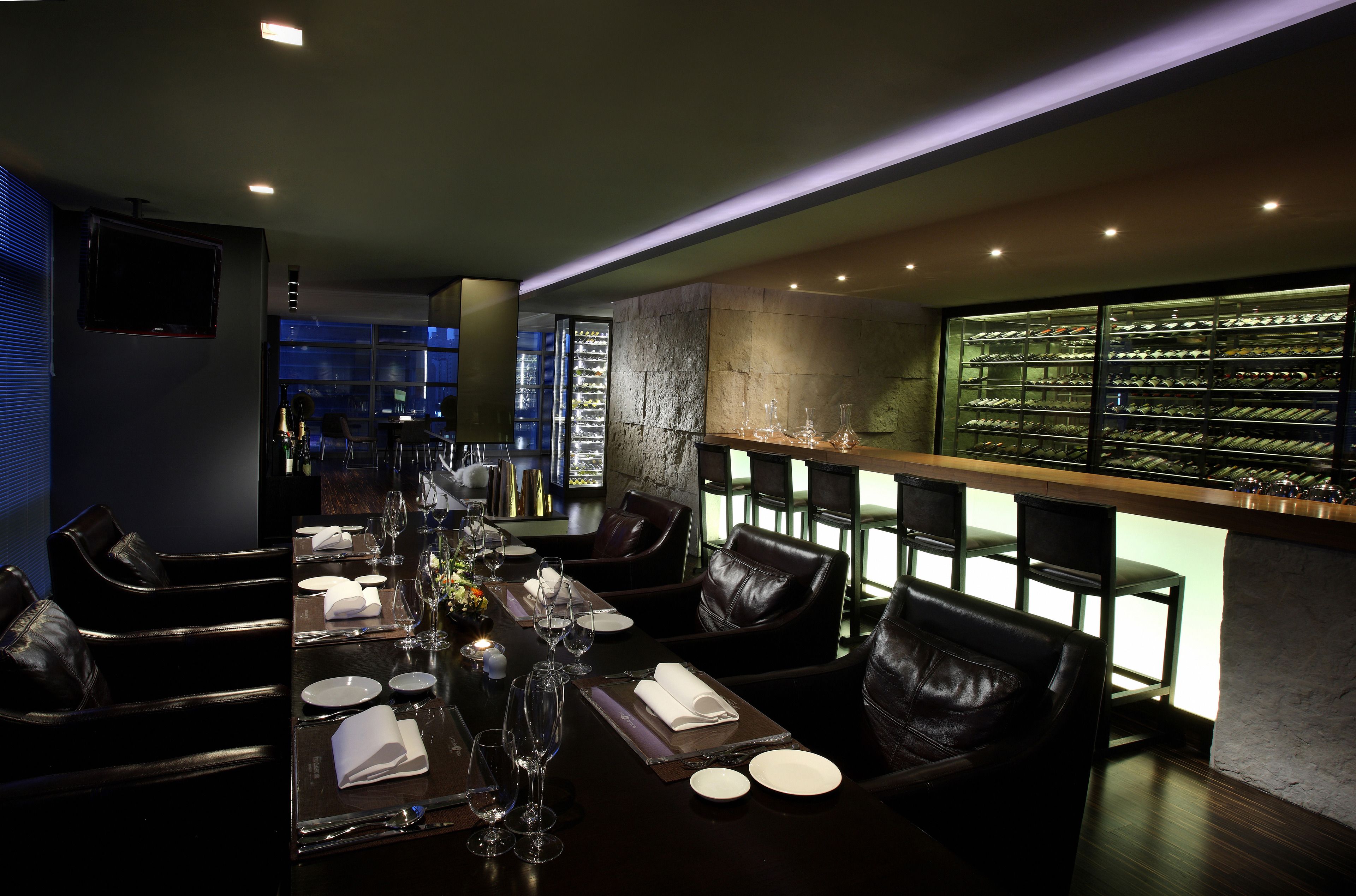 Un restaurante con sillas de cuero oscuro, una larga mesa de comedor dispuesta con platos, copas y cubiertos. Se aprecia una estantería con botellas de vino a la derecha.