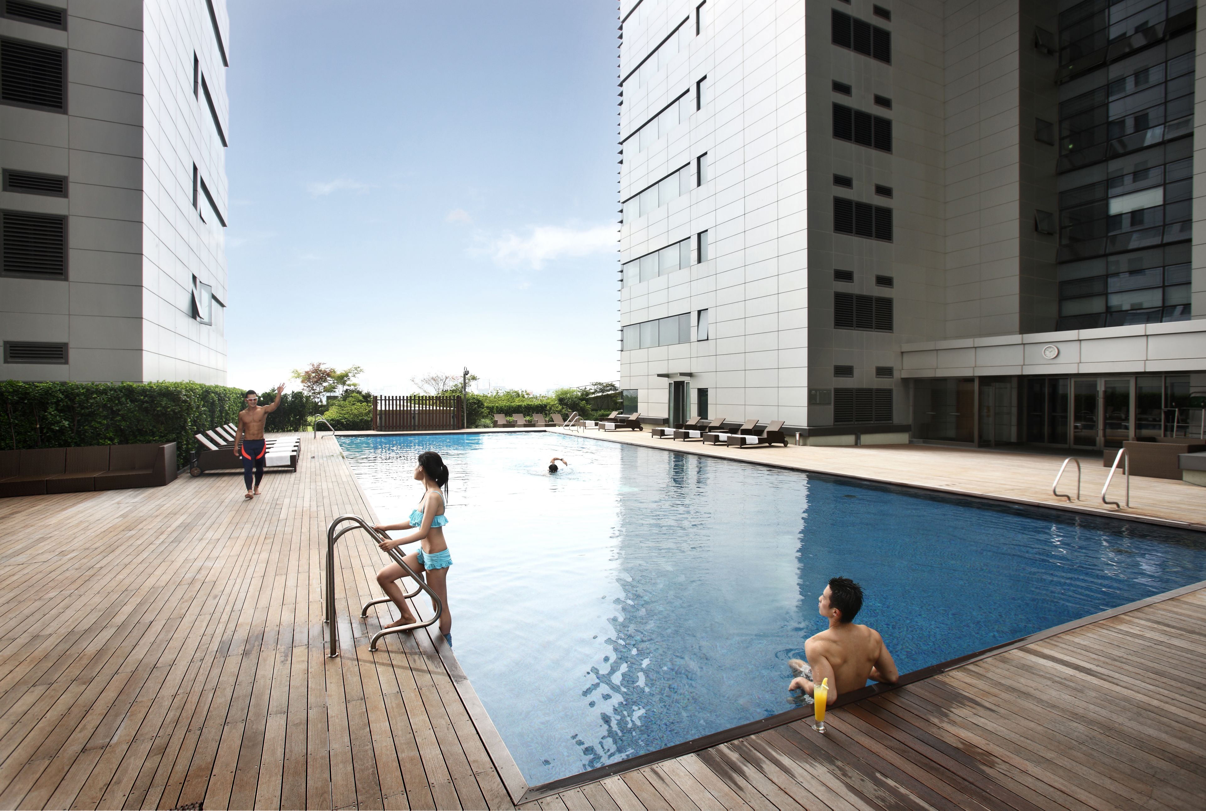 A swimming pool area with a wooden deck, lounge chairs, and a modern building in the background.