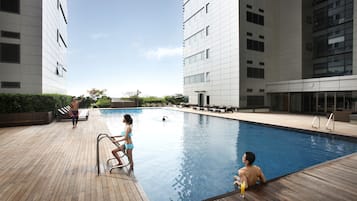 A swimming pool area with a wooden deck, lounge chairs, and a modern building in the background.