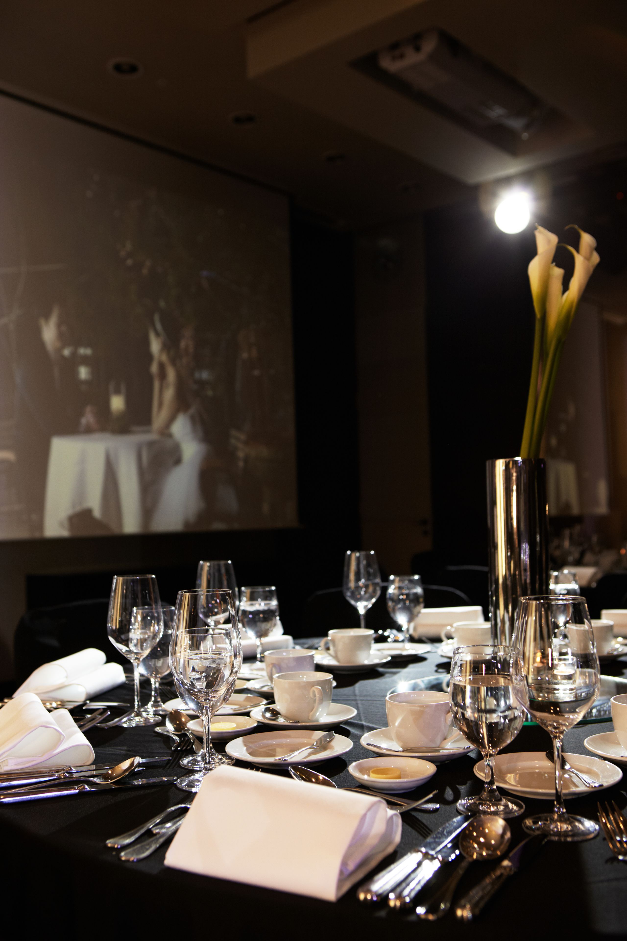 A dining table set with multiple wine glasses, plates, and cutlery, with a projector screen in the background displaying a scene.