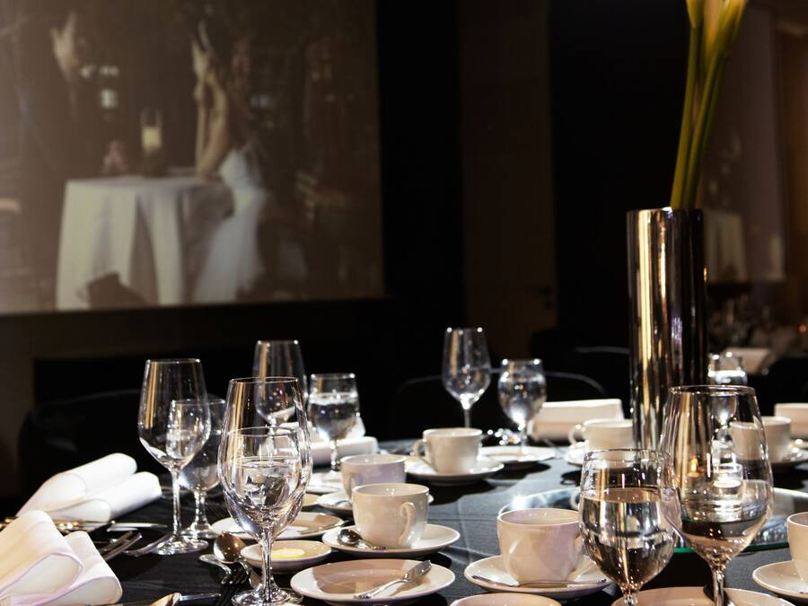 A dining table set with multiple wine glasses, plates, and cutlery, with a projector screen in the background displaying a scene.