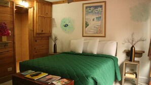 A room with a green quilted bed, wooden walls, a painting, and a small table with books.
