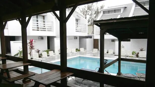 A pool area with wooden tables and chairs, surrounded by white buildings.