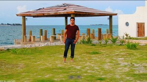 A man standing under a wooden gazebo by a body of water, with a white building and a wooden door in the background.