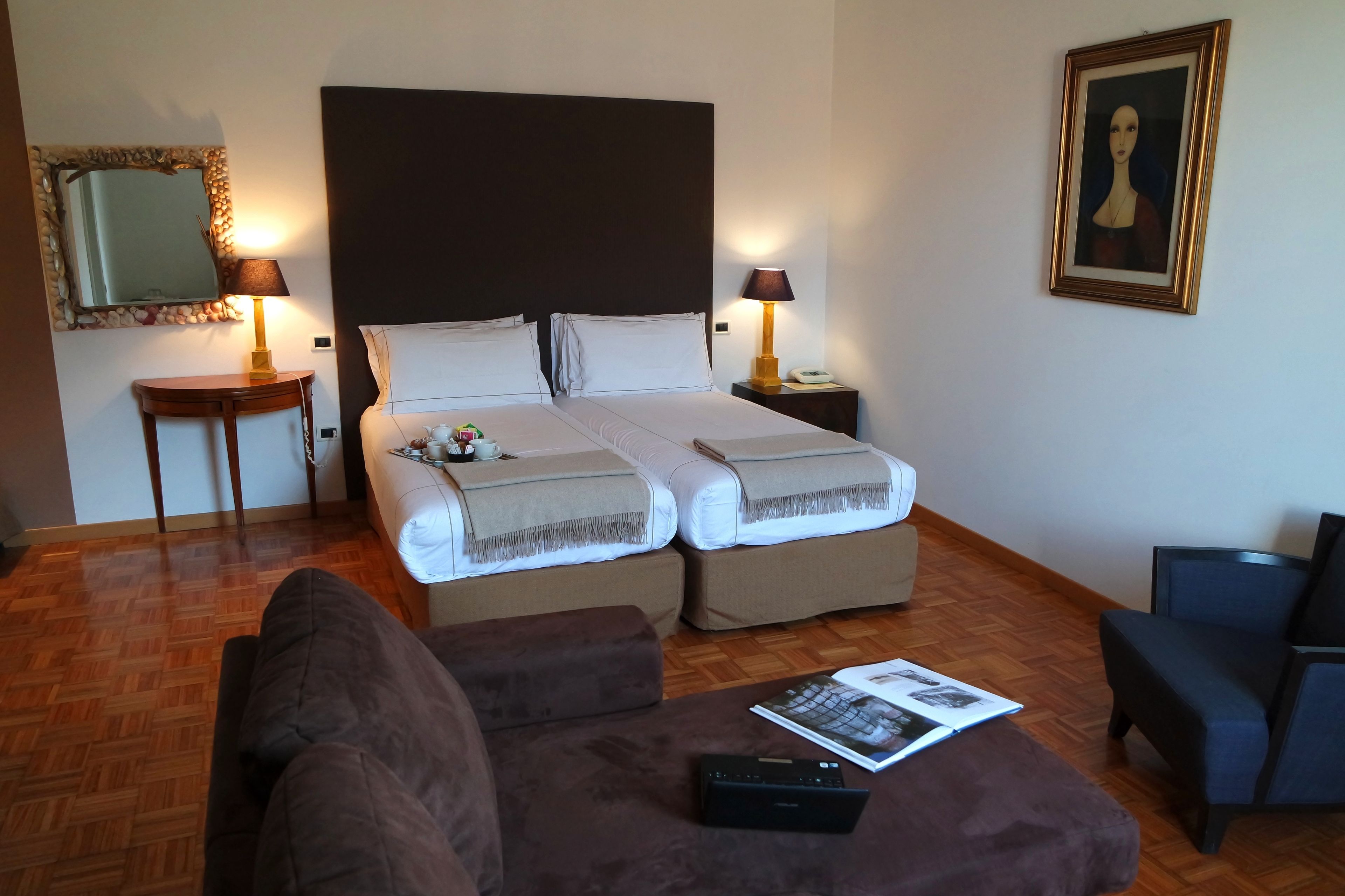 Een hotelkamer met een tweepersoonsbed, twee nachtkastjes, een slaapbank, een fauteuil, een lamp, een ingelijst kunstwerk en een tijdschrift op de poef.