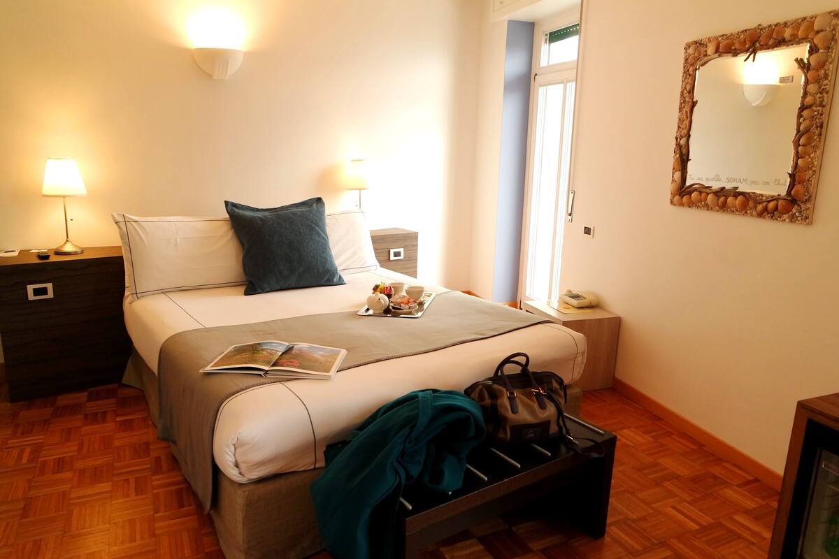 A neatly arranged bedroom with a bed, bedside lamps, a bedside table, a mirror, and a window.