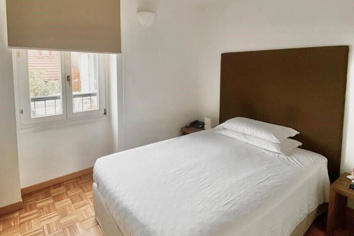 A bedroom with a large bed, a nightstand, a window with a blind, and a wooden floor.