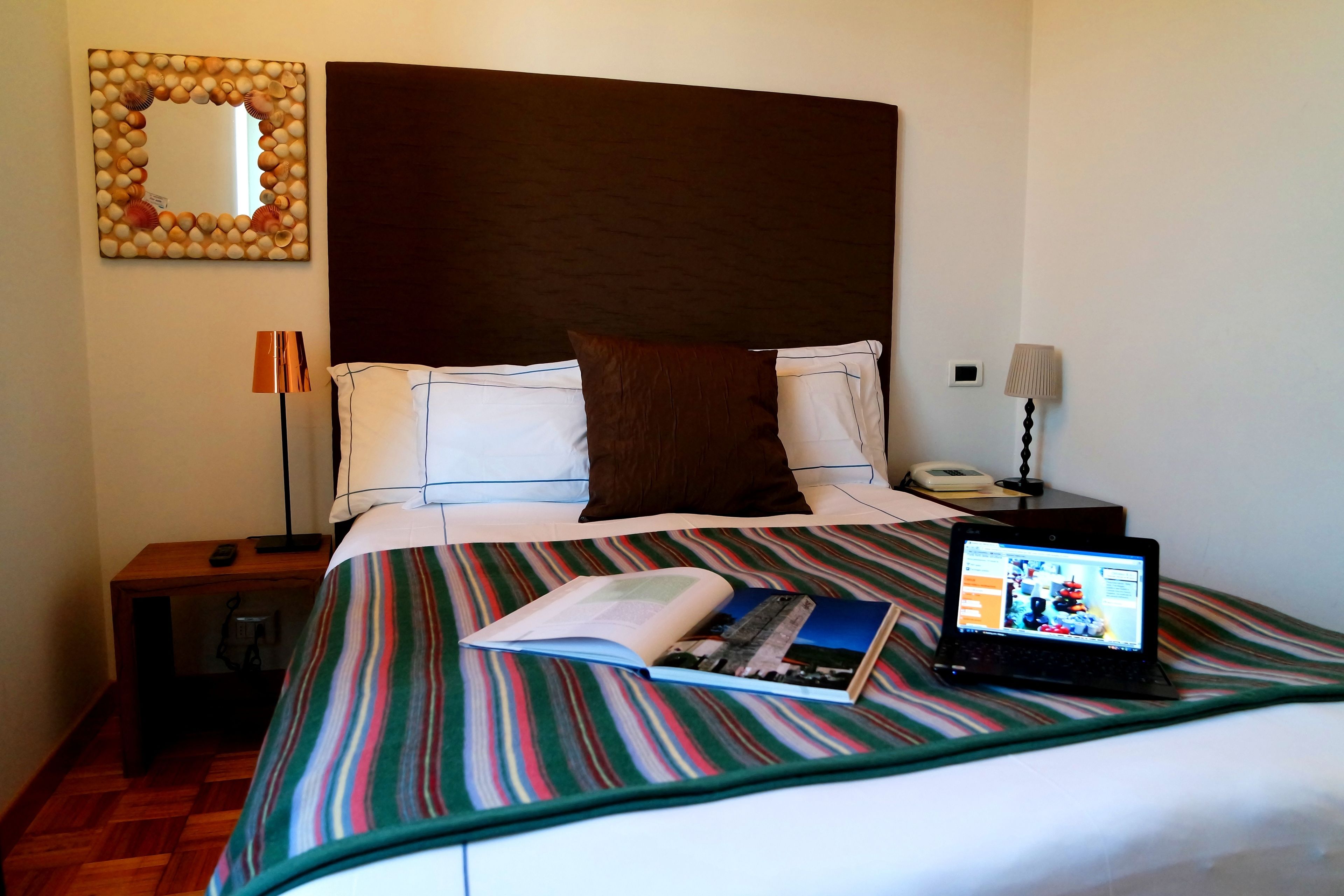 A hotel room with a bed, a laptop, a book, a lamp, and a mirror on the wall.