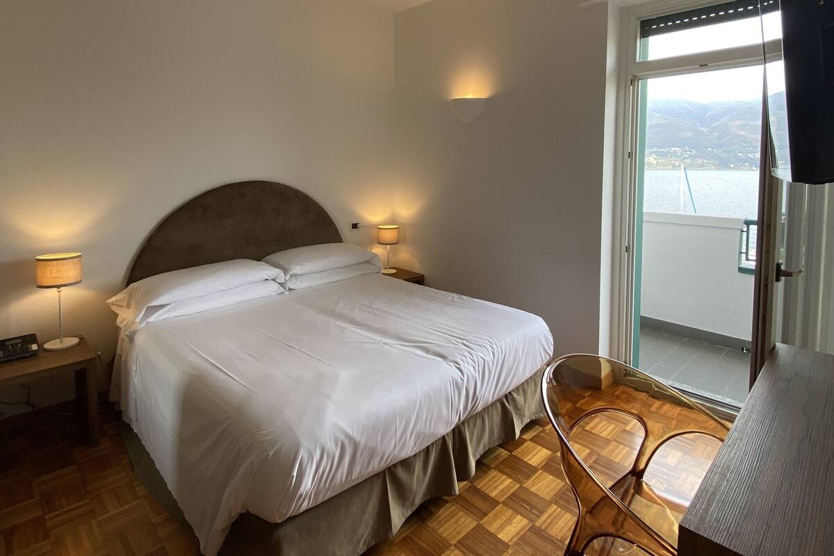 A bedroom with a large bed, two bedside lamps, a wooden floor, a chair, and a view of a body of water through a window.