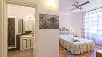 Un dormitorio con cama, mesita de noche, lámpara de pared, ventilador de techo y baño con ducha y lavamanos revestidos de azulejos.