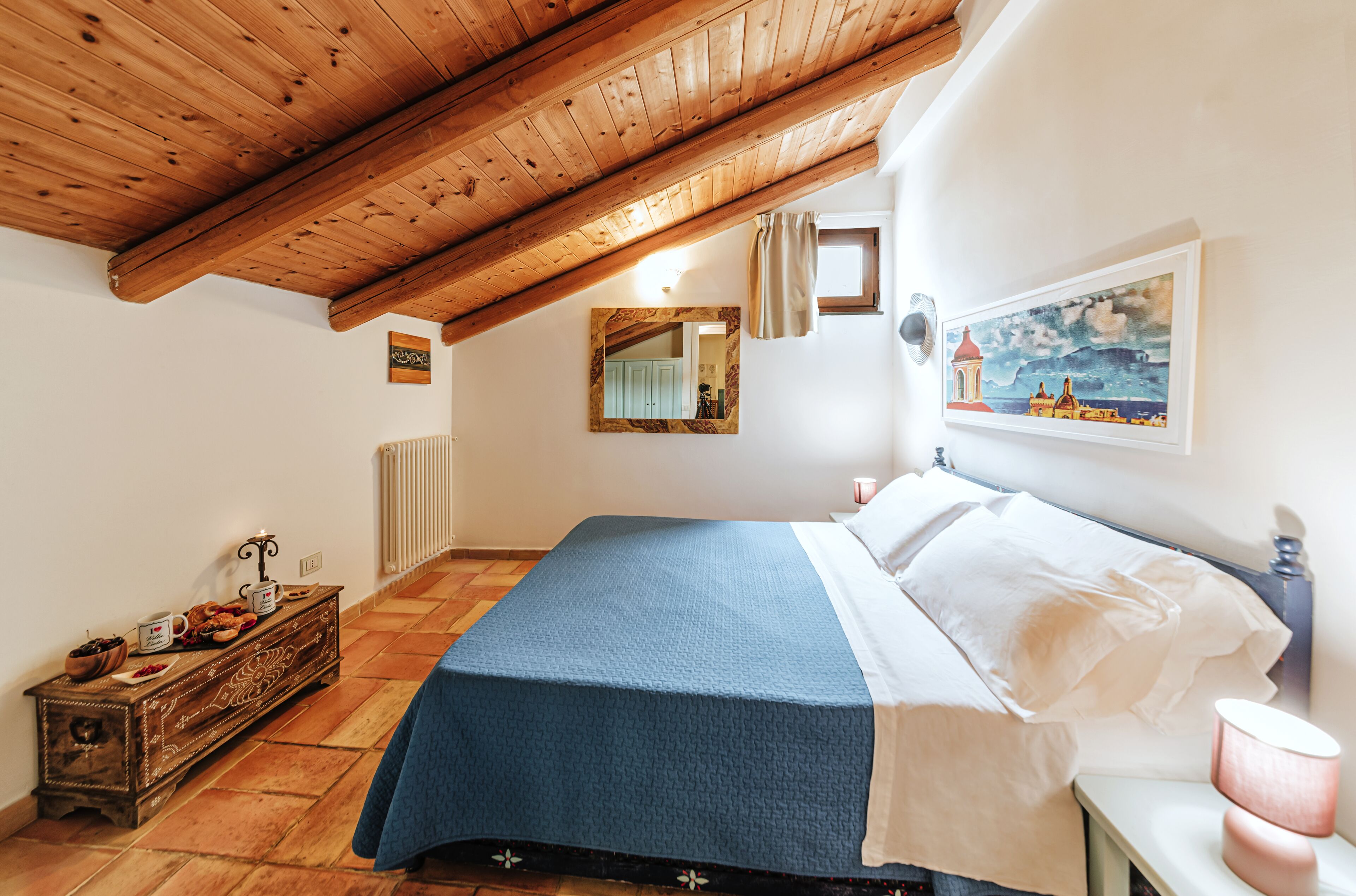 Un dormitorio con techo de madera, una cama con colcha azul, una mesita de noche con lámpara, un aparador con objetos decorativos y un cuadro en la pared.