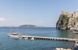 Uma paisagem costeira com um cais que se estende até o mar, barcos atracados no cais e um edifício histórico no topo de um penhasco.