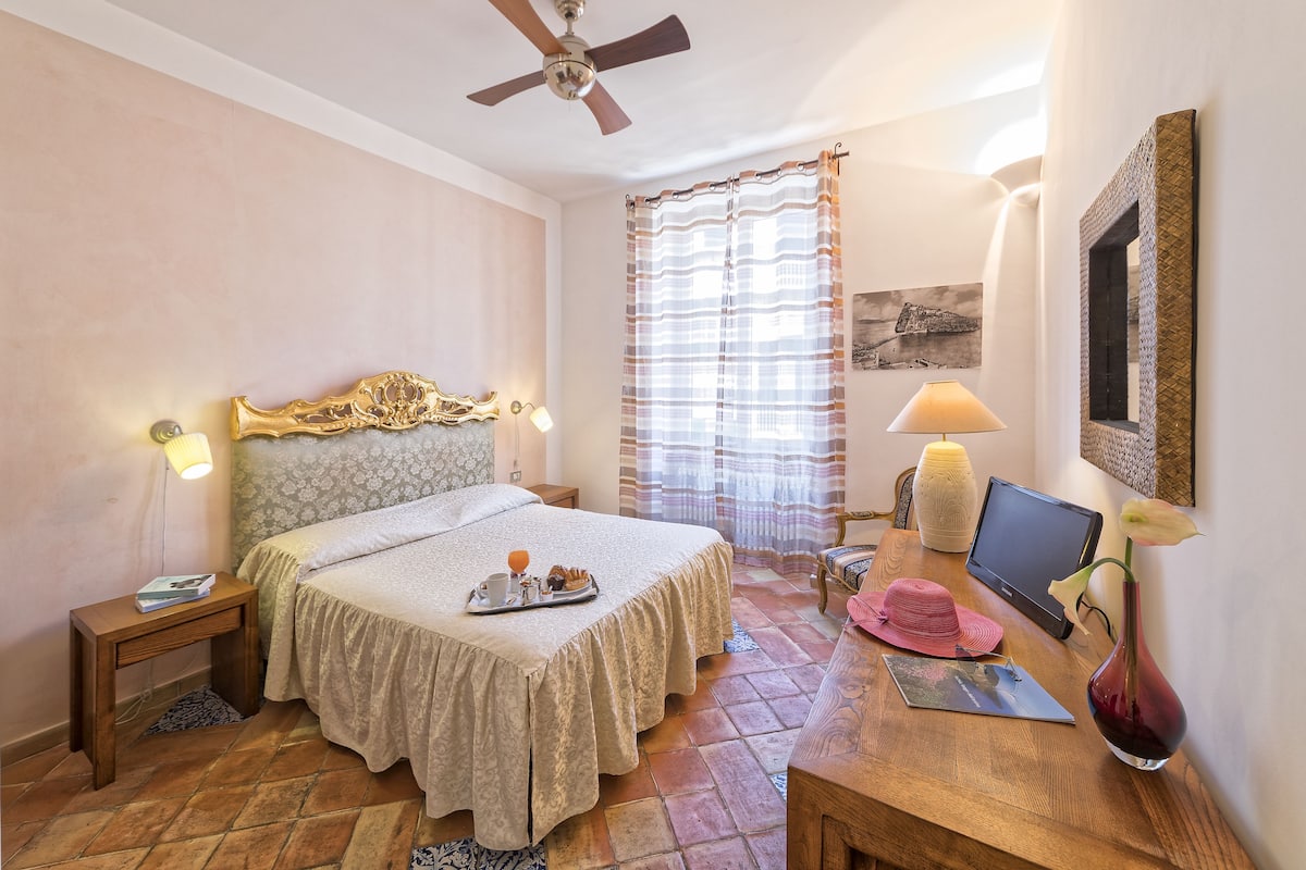 Ein Hotelzimmer mit einem Bett, einem Schreibtisch mit einem Laptop, einer Vase mit einer Blume, einem Sessel und einem Deckenventilator.
