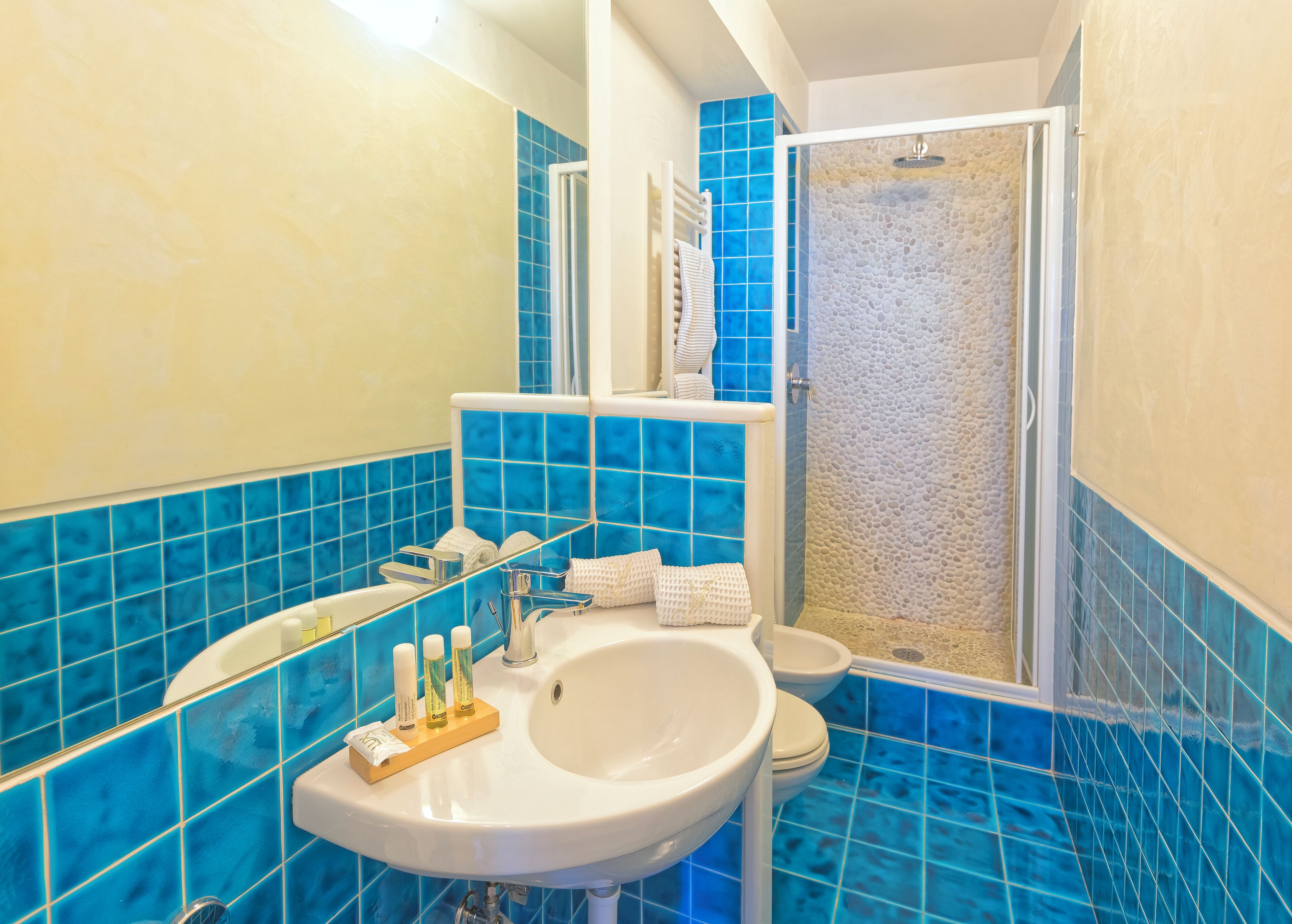 Ein Badezimmer mit einem weißen Waschbecken, blau gefliesten Wänden, einer Dusche mit strukturierter Wand und einem Spiegel über dem Waschbecken.