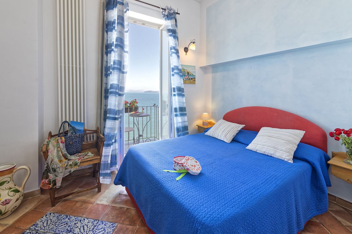 Ein Schlafzimmer mit einem großen Bett, einem Sessel, einem Tisch mit Blumen und Meerblick vom Balkon.
