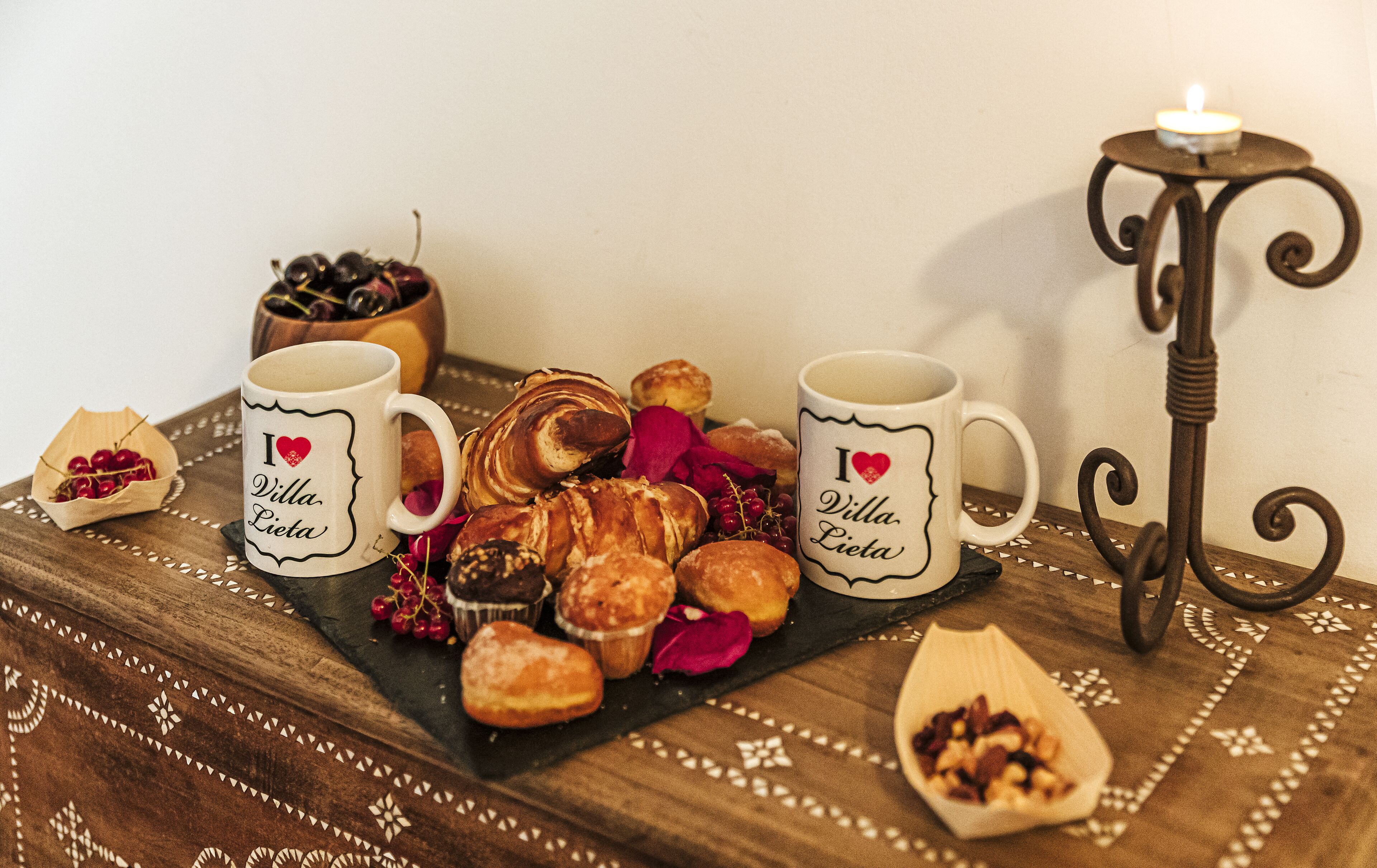 A table with two white mugs, a tray of pastries, a small bowl of cherries, and a candle on a stand.