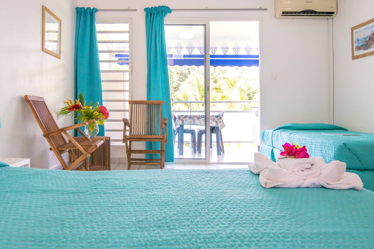 Habitación de hotel con dos camas, una silla de madera, una mesa con sillas, un florero y un balcón con vista a zonas verdes.