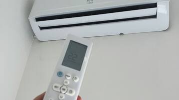 A hand holding a remote control with a digital display, positioned in front of an air conditioning unit.