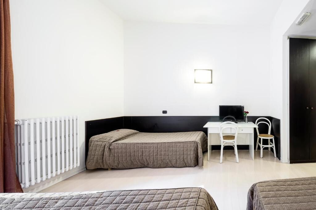 Una habitación pequeña y moderna con una cama, un escritorio y una silla. Hay un radiador en la pared y una puerta a la derecha.