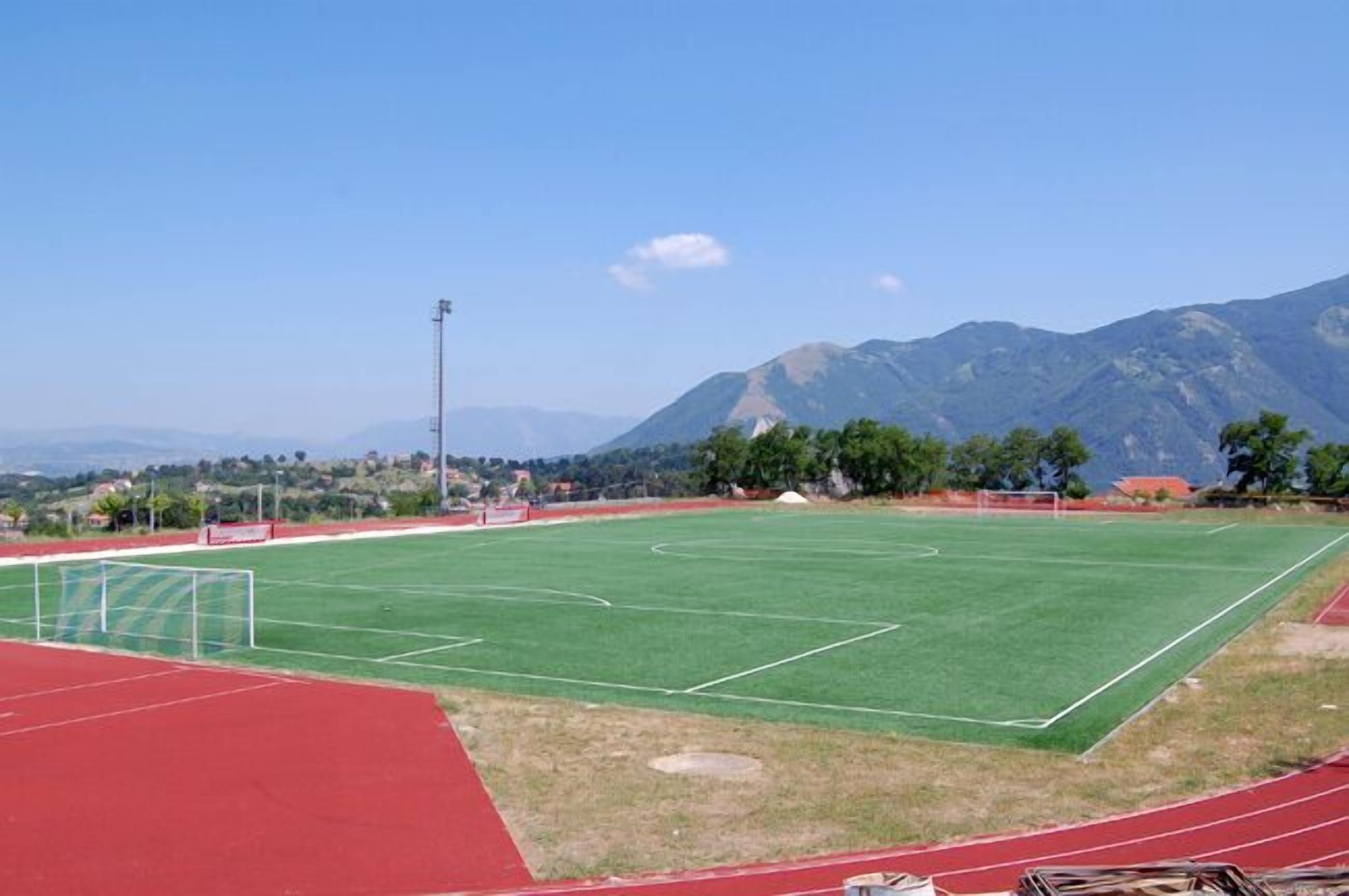 Un campo deportivo con pista de atletismo y postes de portería, rodeado de montañas.