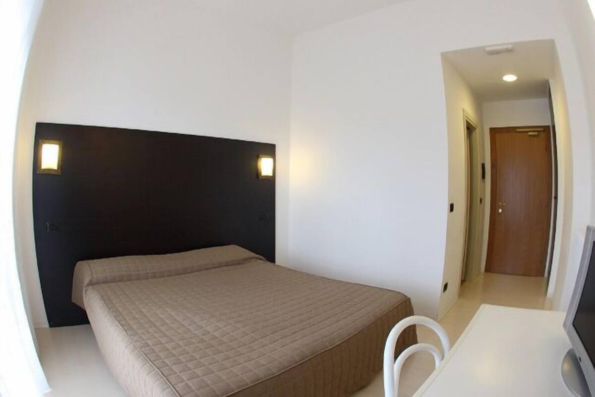 A bedroom with a bed, a desk, and a chair. There is a door and a light fixture on the wall.