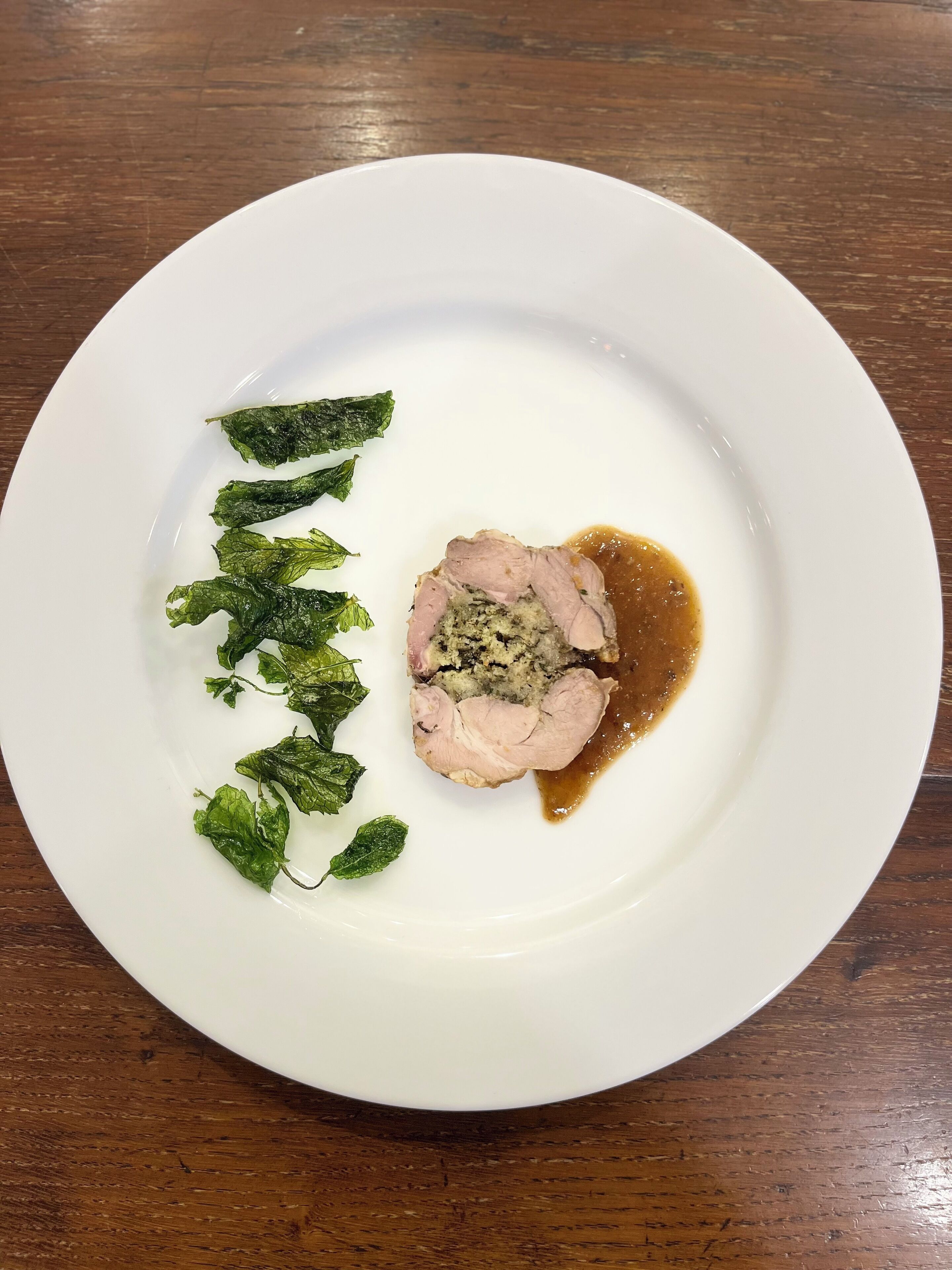 A white plate with a slice of meat, possibly pork, and a brown sauce, accompanied by green leaves on a wooden table.