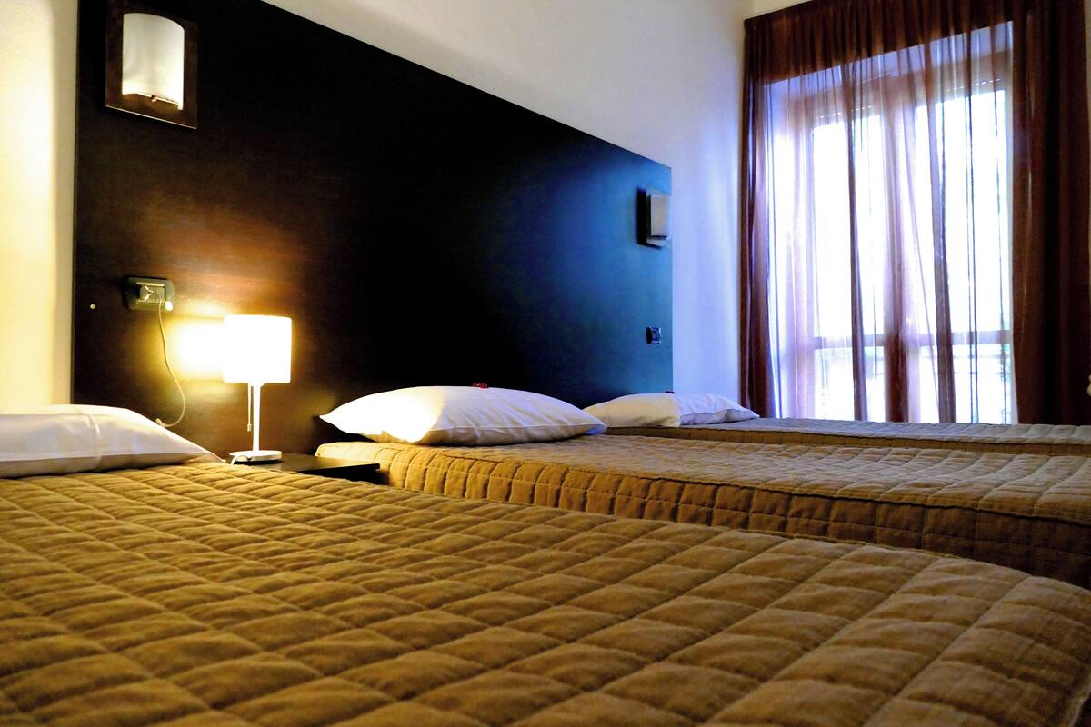 A hotel room with two beds, a nightstand with a lamp, and a window with curtains.