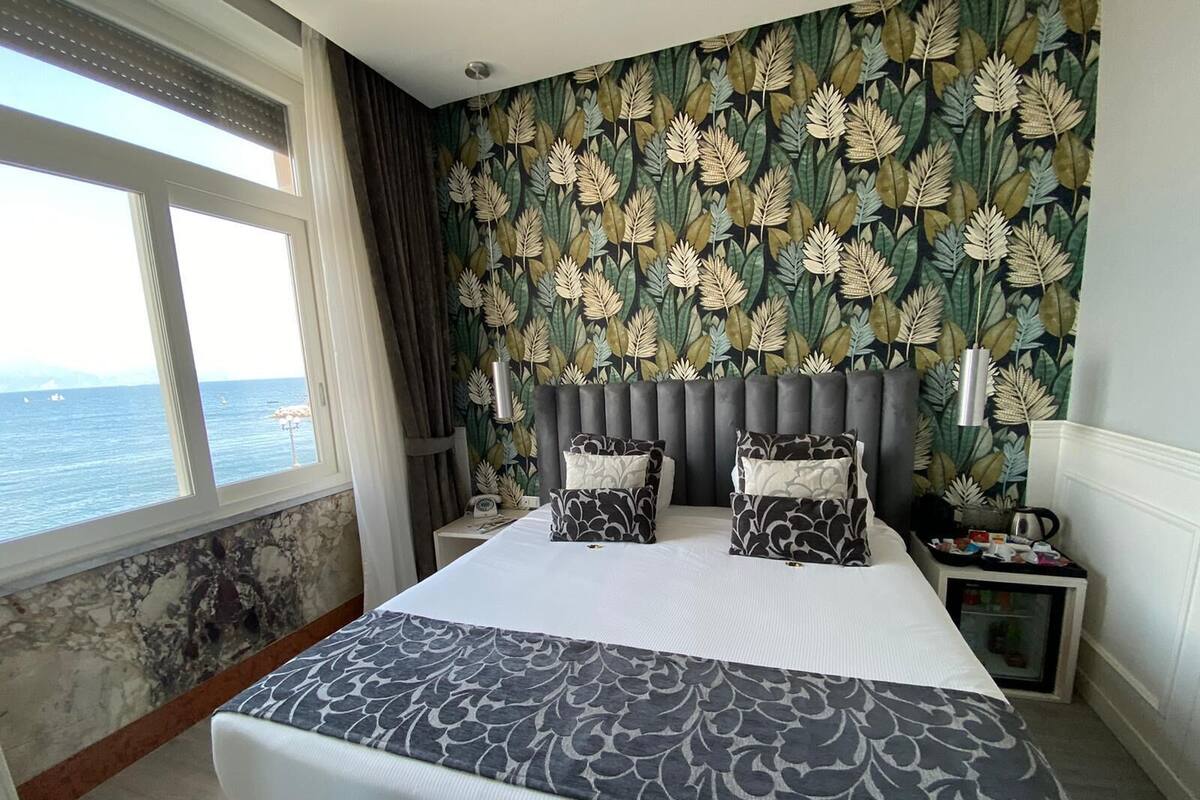 Une chambre d’hôtel avec un grand lit, une vue sur la mer, un papier peint à motifs et une table de chevet avec une lampe.