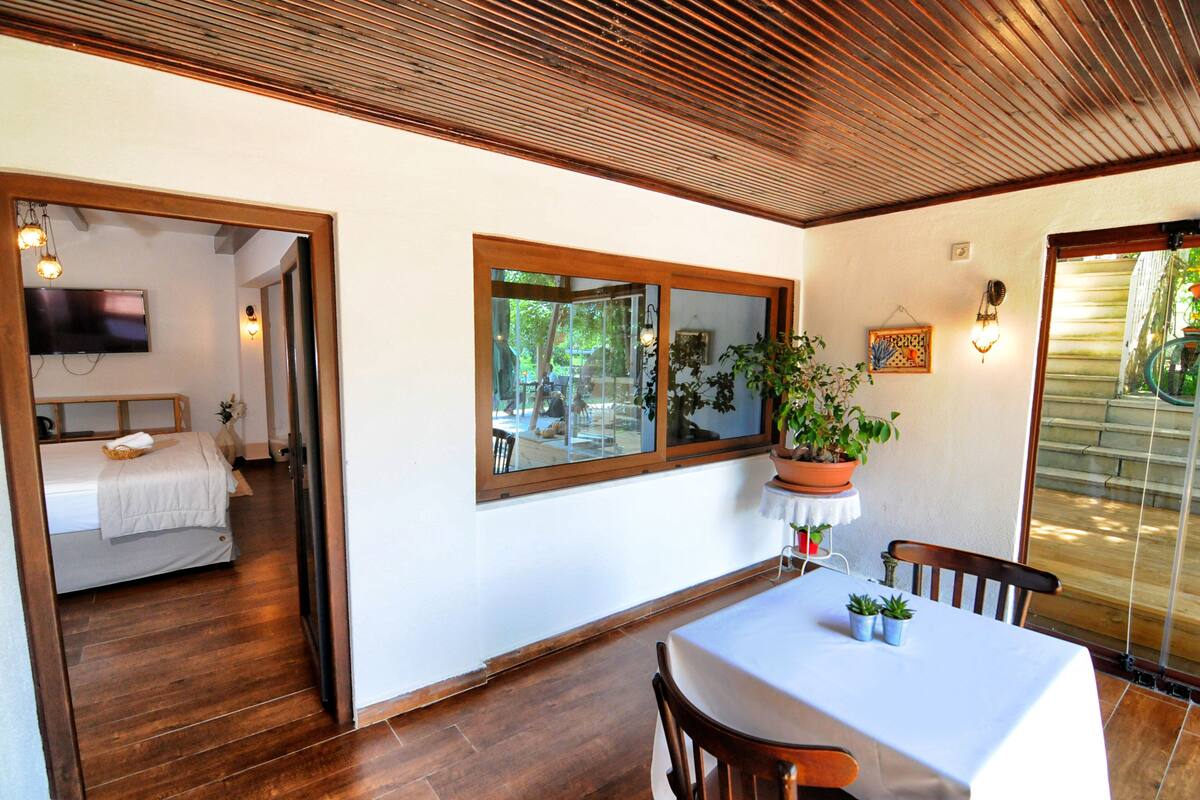 A room with a wooden ceiling, a large window with a view, a bed, a table with chairs, and a potted plant.