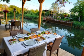 Una mesa de comedor lista para una comida, con varios platos, rodeada de sillas de mimbre y con vistas a una masa de agua tranquila, una terraza de madera y una barandilla de cadena.