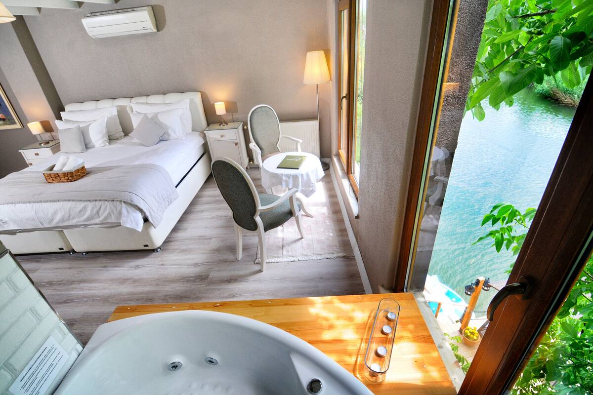 A hotel room with a bed, a chair, a small table, a bathtub, and a view of a body of water.