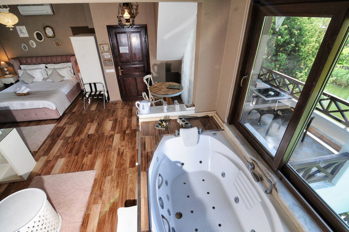 A bedroom with a large bed, a wooden floor, a small table with a kettle, and a window with a view of greenery.