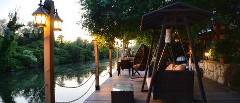 A wooden deck with a swing chair, a table, and chairs, overlooking a calm body of water.