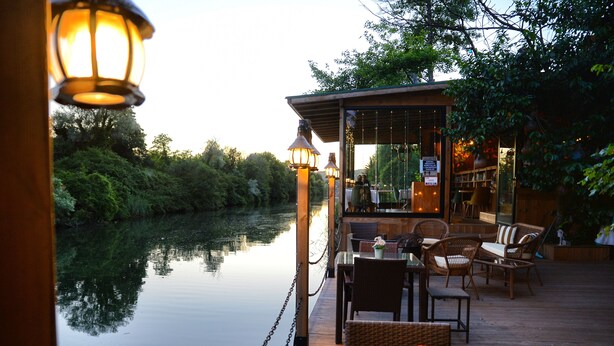 Una terraza tranquila junto al río, con mobiliario de mimbre y un pequeño edificio, presumiblemente un restaurante o bar, que ofrece vistas al agua y al follaje.