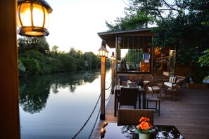Una terraza tranquila junto al río, con mobiliario de mimbre y un pequeño edificio, presumiblemente un restaurante o bar, que ofrece vistas al agua y al follaje.