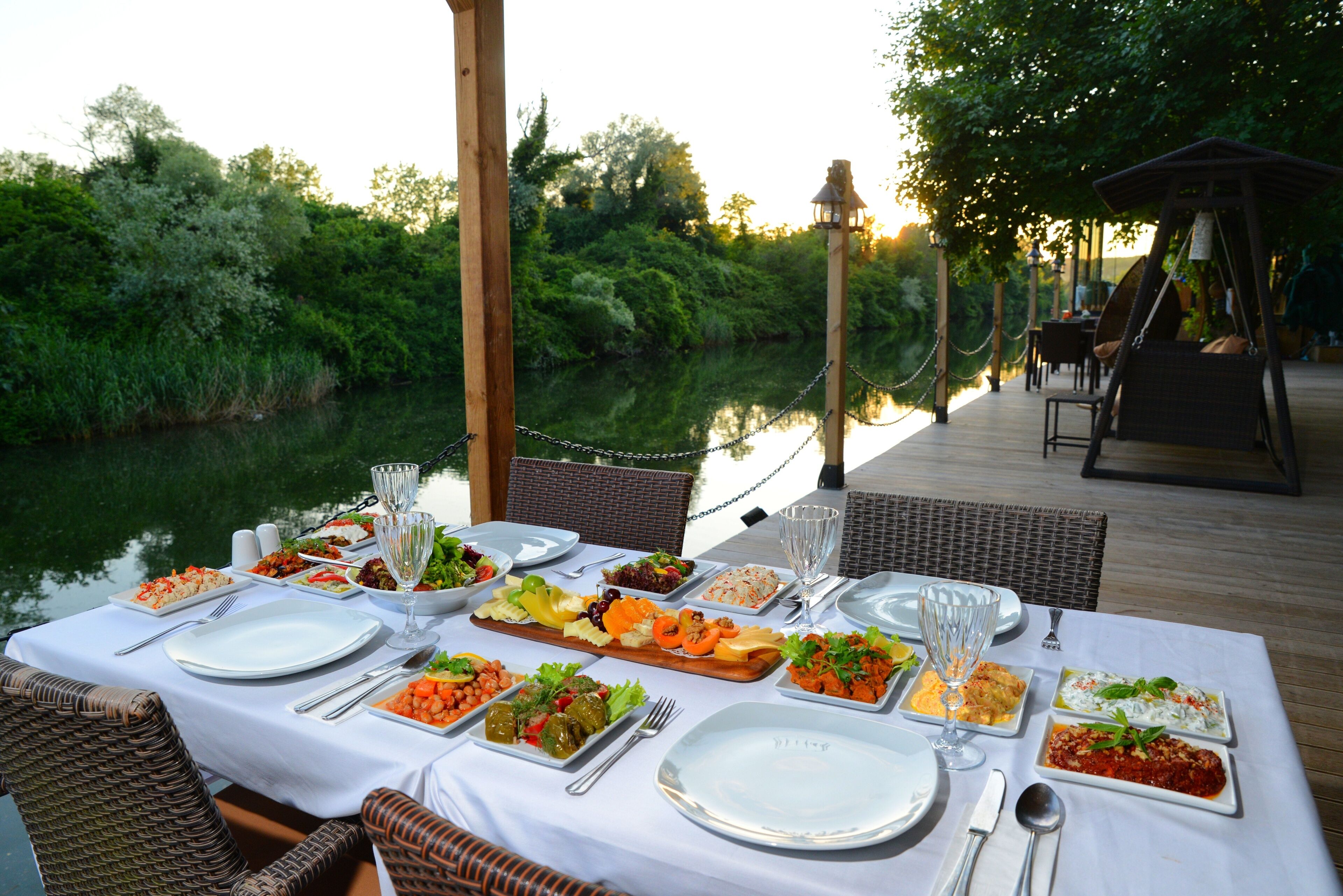 An outdoor dining area with a table set for a meal, surrounded by wicker chairs and a view of a river.