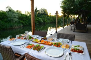 Un espacio de comedor al aire libre con una mesa dispuesta para una comida, rodeado de sillas de mimbre y vistas a un río.