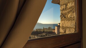 Blick aus einem Fenster mit Steinwand und Vorhang auf eine Küstenstadt mit Gebäuden und blauem Himmel.