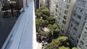Un balcón con una mesa y sillas de vidrio, con vistas a un paisaje urbano con rascacielos y una calle arbolada.