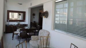 Un comedor con mesa de vidrio, sillas de madera y un amplio ventanal con persianas.