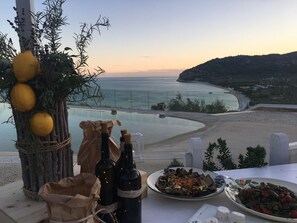 Une table avec des bouteilles, une assiette d’olives et une vue magnifique sur la mer au coucher du soleil.