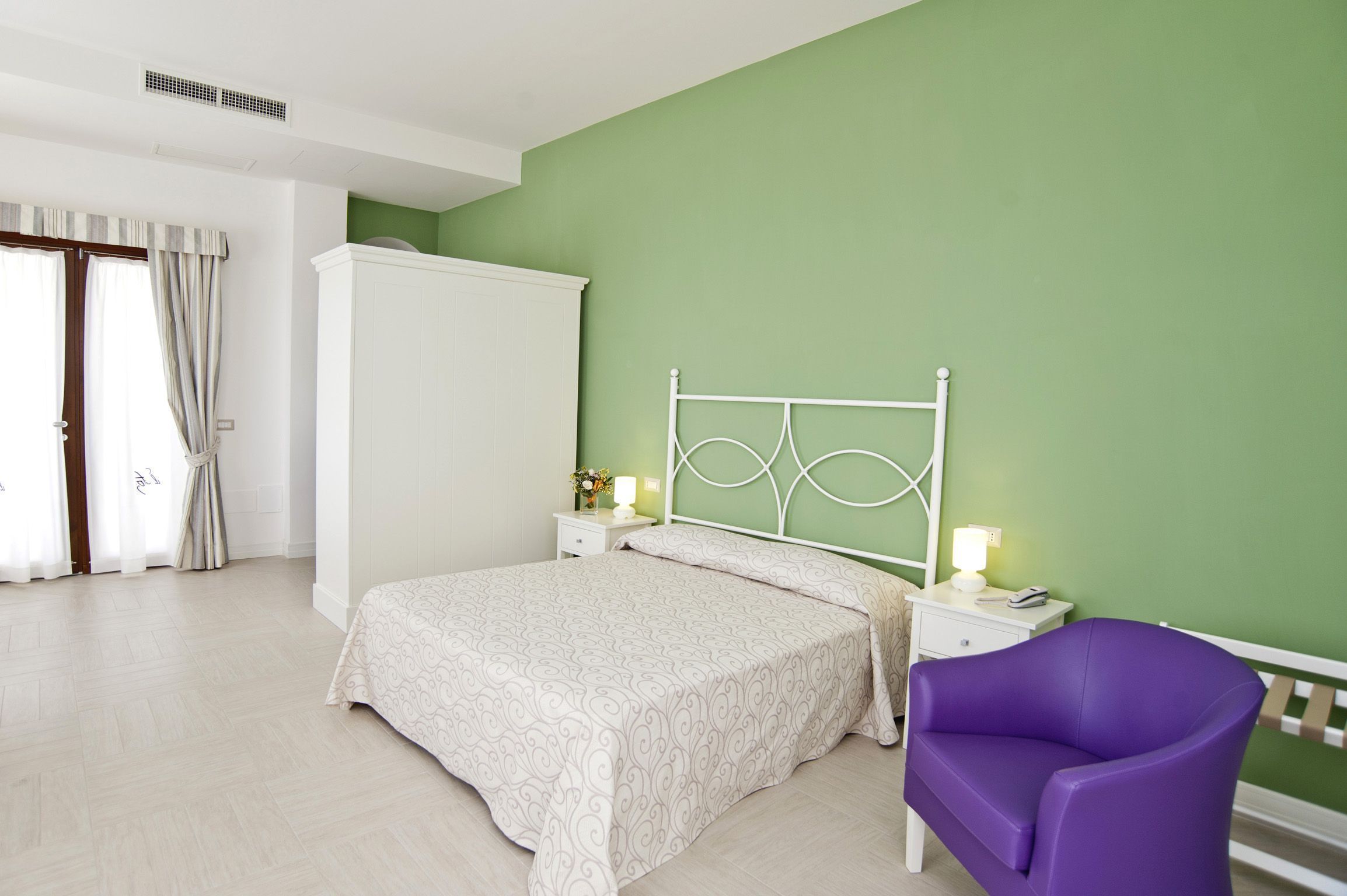 Un dormitorio con cama, mesita de noche, silla, armario y ventana con cortinas.