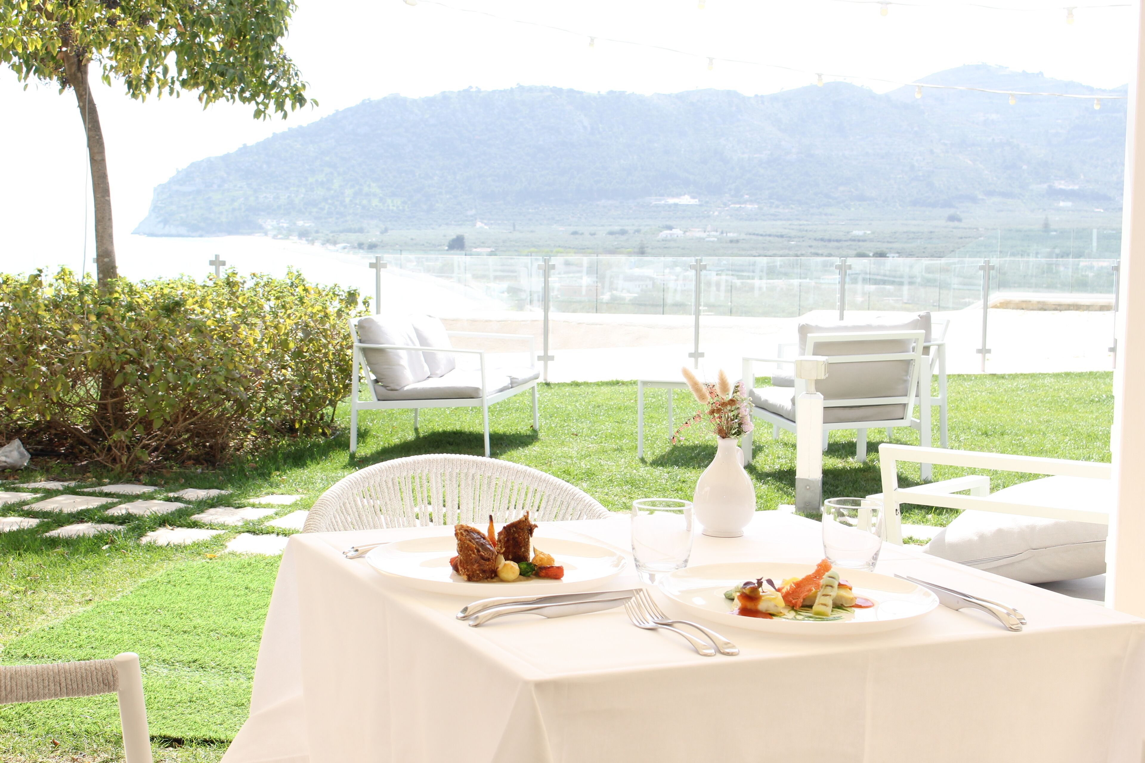 An outdoor dining area with white furniture, a table set with plates of food, and a view of a body of water and hills in the background.