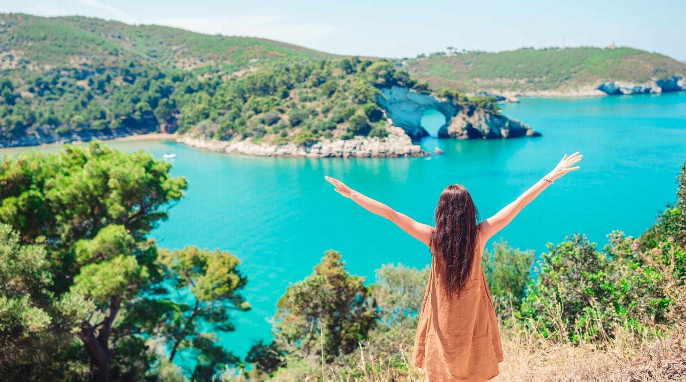A person with arms outstretched overlooking a coastal landscape with trees and a clear sky.